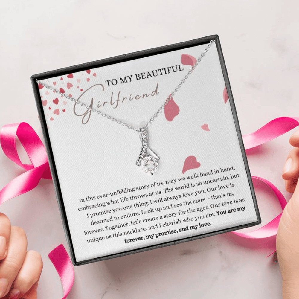 A person unwrapping a necklace gift with ribbon shaped pendant with cubic zirconia crystals and white gold finish, with a message card to my beautiful girlfriend.