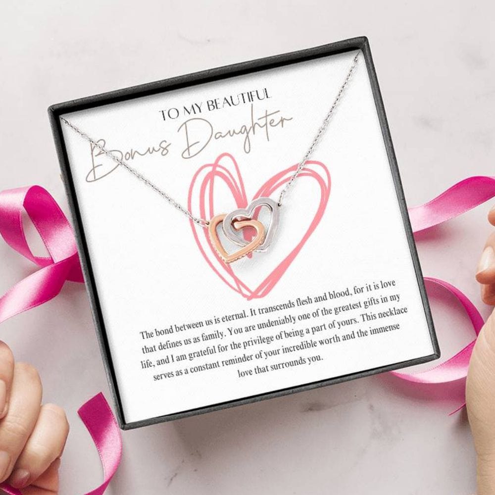 A person unwrapping a necklace gift, with two connected hearts embellished with cubic zirconia crystals and white gold finish, with a message card, to my beautiful bonus daughter.