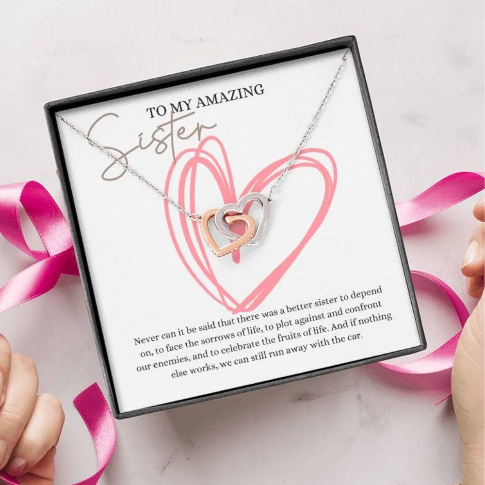 A person unwrapping a necklace gift, with two connected hearts embellished with cubic zirconia crystals and white gold finish, with a message card, to my amazing sister.