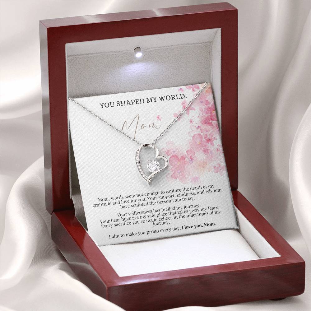 A white gold finish necklace gift, featuring a stunning 6.5mm CZ crystal surrounded by a polished heart pendant embellished with smaller crystals, with a message card to mom, next to a mahogany Jewelry box