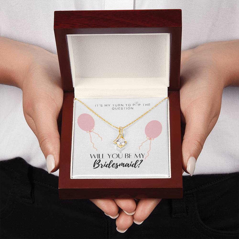 A woman presenting a mahogany Jewelry gift box with a necklace with ribbon shaped pendant made from cubic zirconia crystals and a gold finish, with a message card to bridesmaids.