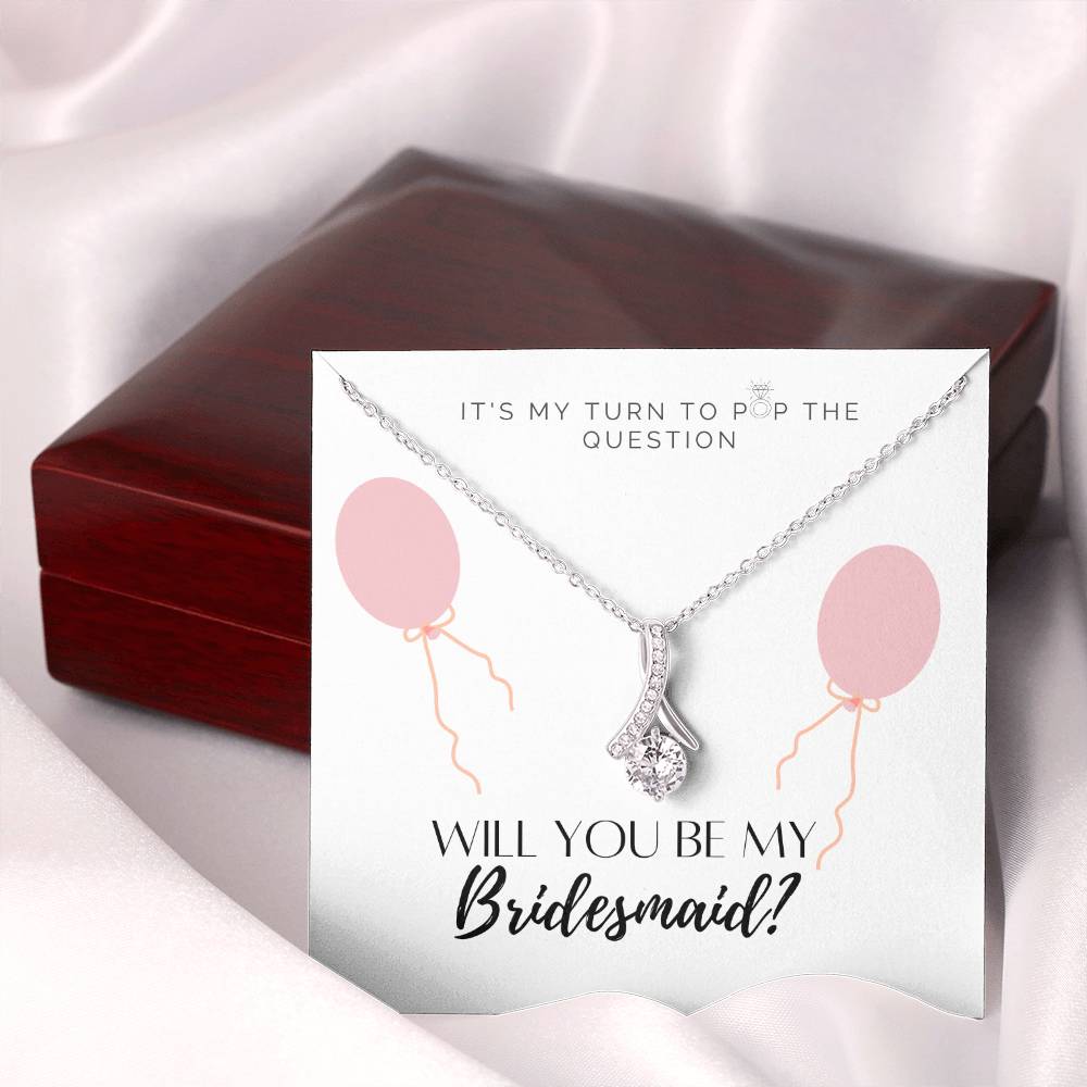 A necklace gift with ribbon shaped pendant with cubic zirconia crystals and white gold finish, with a message card to bridesmaids next to a mahogany Jewelry box.