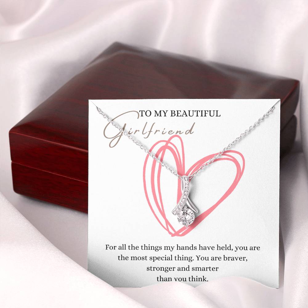 A necklace gift with ribbon shaped pendant with cubic zirconia crystals and white gold finish, with a message card to my beautiful girlfriend next to a mahogany Jewelry box.