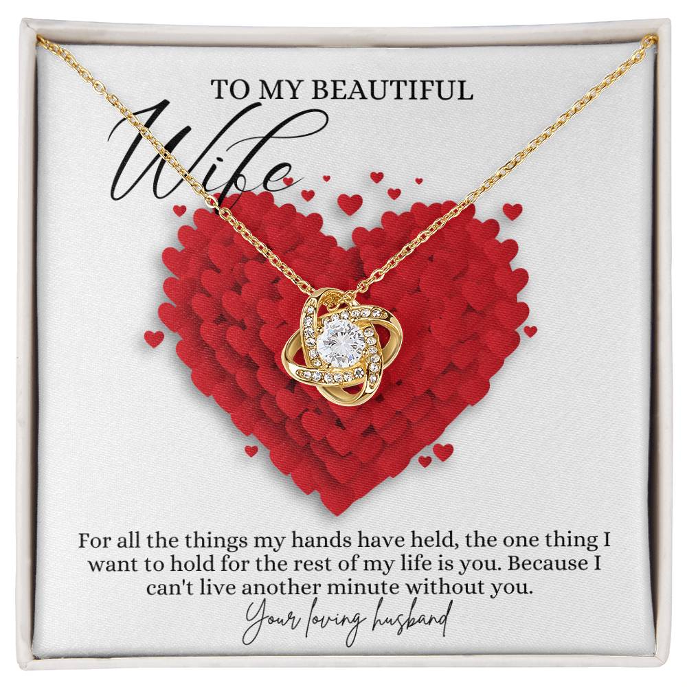 A yellow gold finish necklace gift, with a knot pendant embellished with premium cubic zirconia crystals, and a message card to my beautiful wife, next to a mahogany.