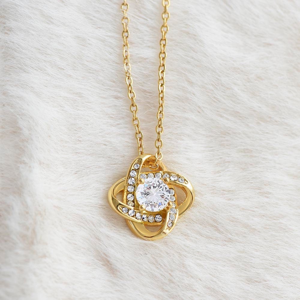 A yellow gold finish necklace gift, with a knot pendant embellished with premium cubic zirconia crystals, laying on a white furry carpet.