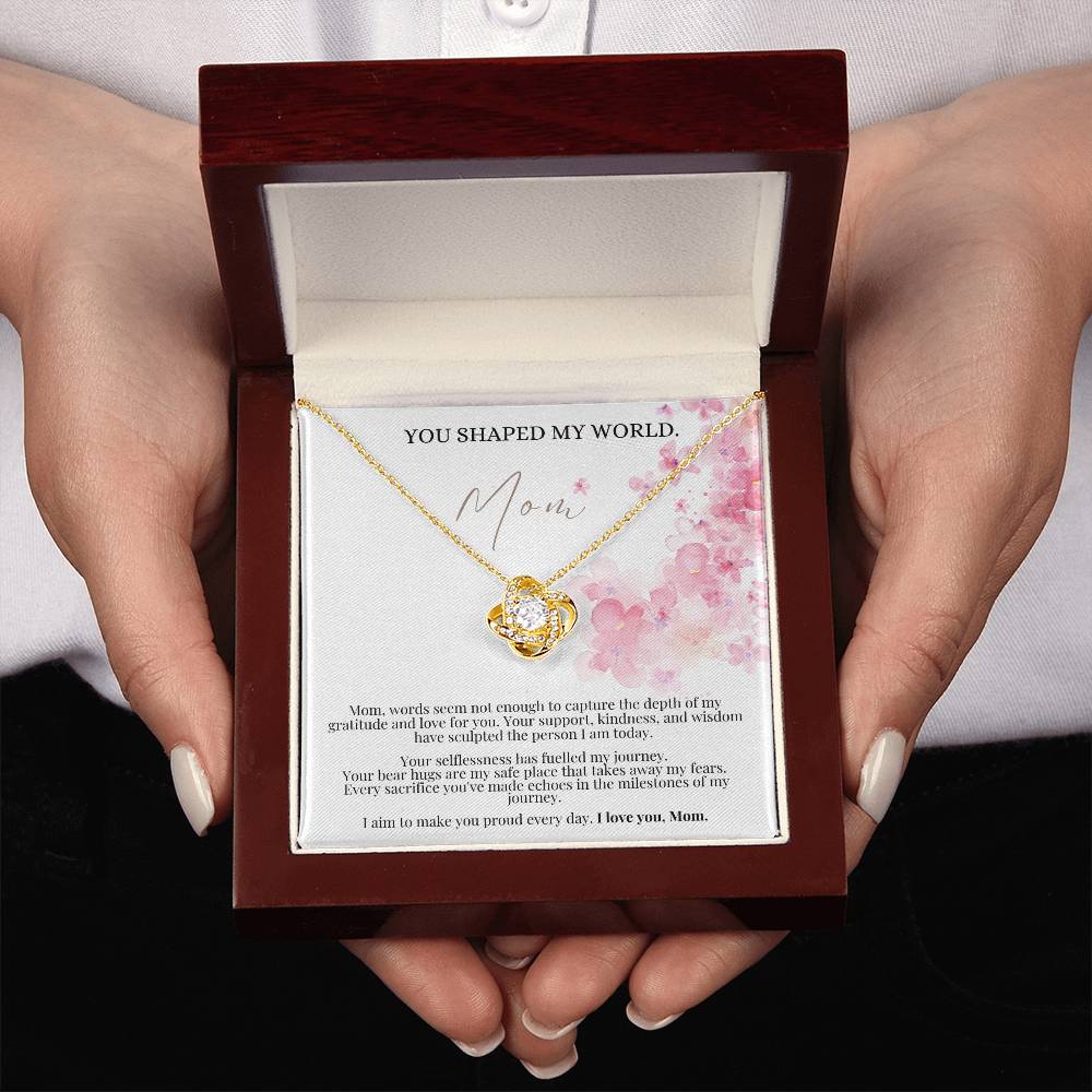 A woman presenting a jewelry box, a yellow gold finish necklace gift, with a knot pendant embellished with premium cubic zirconia crystals, and a message card to mom.