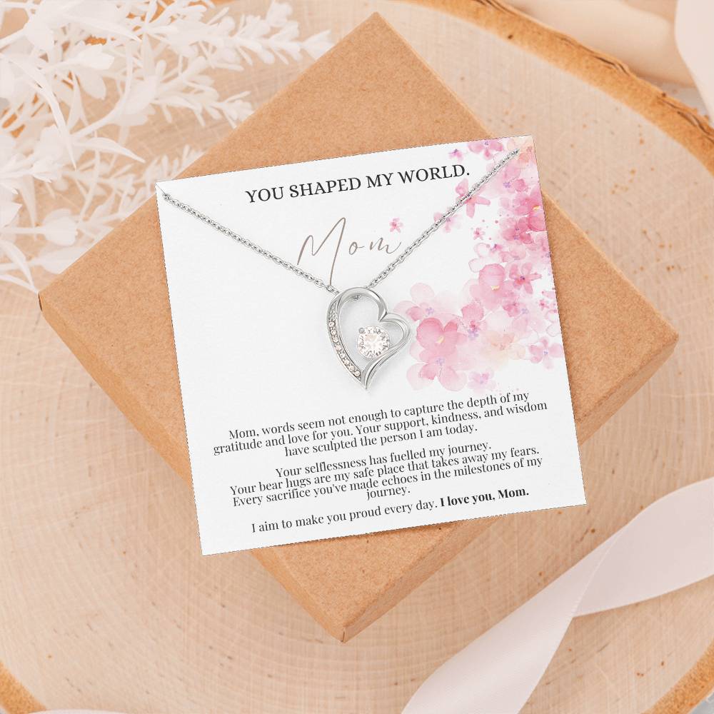 A white gold finish necklace gift, featuring a stunning 6.5mm CZ crystal surrounded by a polished heart pendant embellished with smaller crystals, with a message card to mom, laying on top of a Jewelry box.