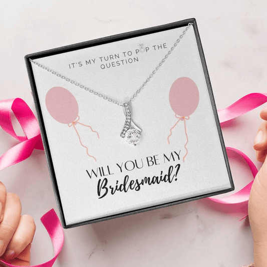 A person unwrapping a necklace gift with ribbon shaped pendant with cubic zirconia crystals and white gold finish, with a message card to bridesmaids. 